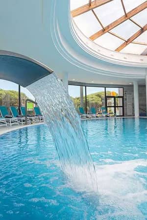 Indoor pool area at Barosa Park featuring a waterfall, sun loungers, and a bright skylight ceiling.