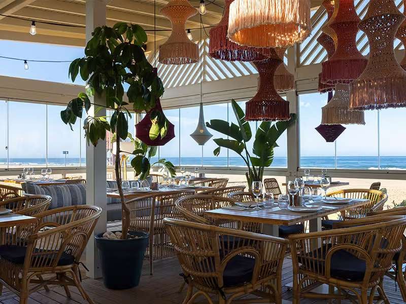 Elegant seaside outdoor dining area at Barosa Park Gallery, with rattan chairs, woven light fixtures, and ocean views through glass walls.