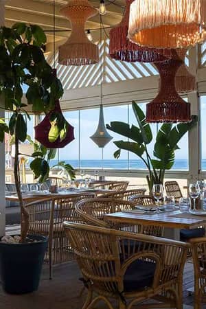 Elegant seaside outdoor dining area at Barosa Park Gallery, with rattan chairs, woven light fixtures, and ocean views through glass walls.