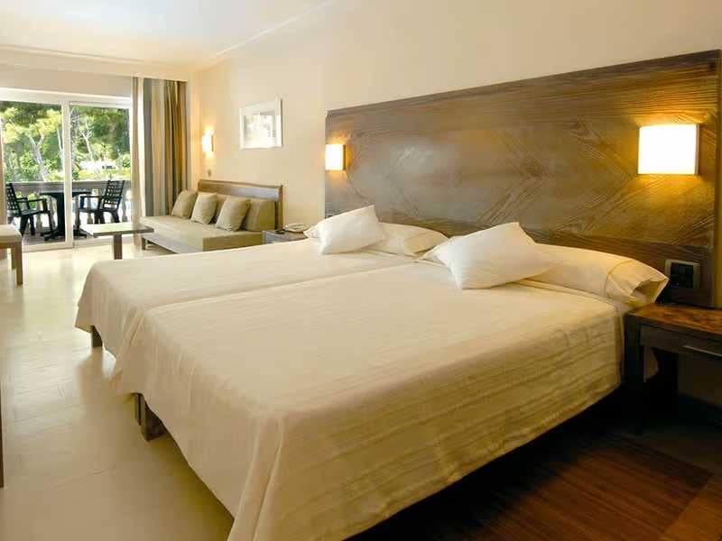 Hotel room with twin beds, seating area, and glass doors opening to a balcony with green views.