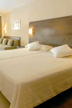 Hotel room with twin beds, seating area, and glass doors opening to a balcony with green views.
