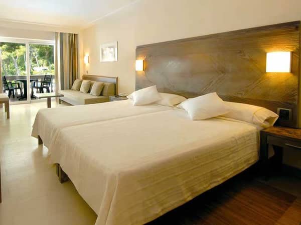 Hotel room with twin beds, seating area, and glass doors opening to a balcony with green views.