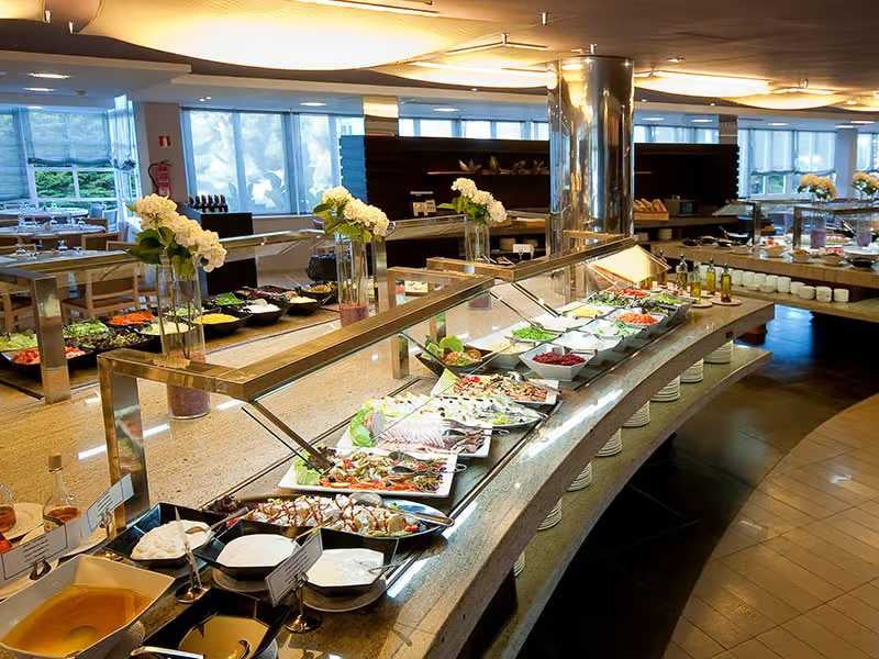 Indoor buffet spread at a beach club with a variety of dishes, salads, platters, and desserts, with decorative flowers and natural light from large windows.