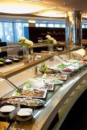 Indoor buffet spread at a beach club with a variety of dishes, salads, platters, and desserts, with decorative flowers and natural light from large windows.