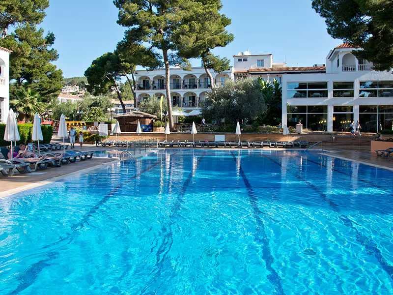 Pool and lounge area at a beach club with sun loungers, parasols, tall trees, and modern white buildings in the background.