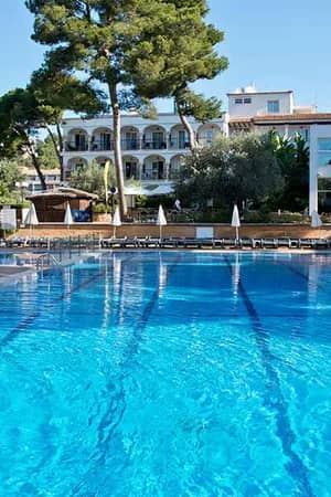 Pool and lounge area at a beach club with sun loungers, parasols, tall trees, and modern white buildings in the background.