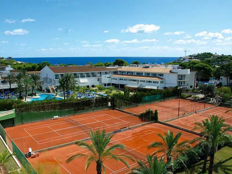 Aerial view of a beach club with clay tennis courts, a pool, white buildings, and sea in the background.