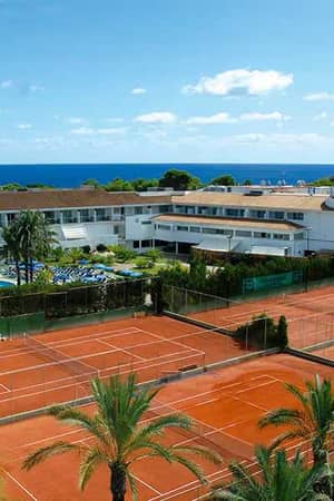 Aerial view of a beach club with clay tennis courts, a pool, white buildings, and sea in the background.