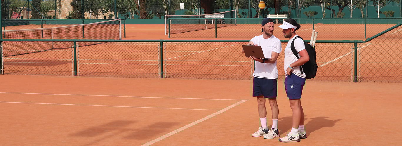Two men on a clay tennis court discussing game data with a laptop in hand.