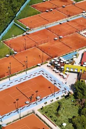 Aerial view of a tennis complex with several red clay courts, surrounded by greenery and parking facilities.