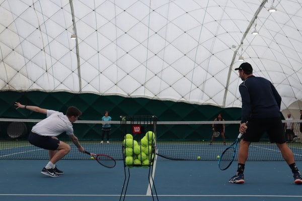 Tennis coaches on an indoor bubble court, practising with a basket of tennis balls in the foreground.