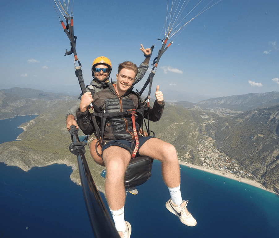 Two men paragliding over a coastal area with blue waters and green mountains, one man smiling and giving thumbs up.