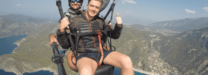 Two men paragliding over a coastal area with blue waters and green mountains, one man smiling and giving thumbs up.