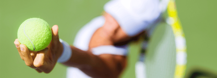 Close-up of a tennis player serving on a green court, focusing on their hand holding a tennis ball and their racket in the background.