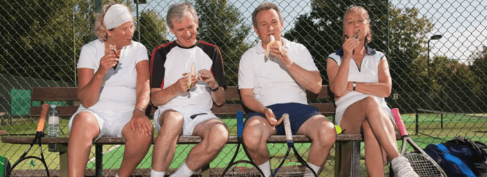 Four adults in tennis gear sitting on a bench by a tennis court, enjoying ice cream cones with rackets and a sports bag nearby.