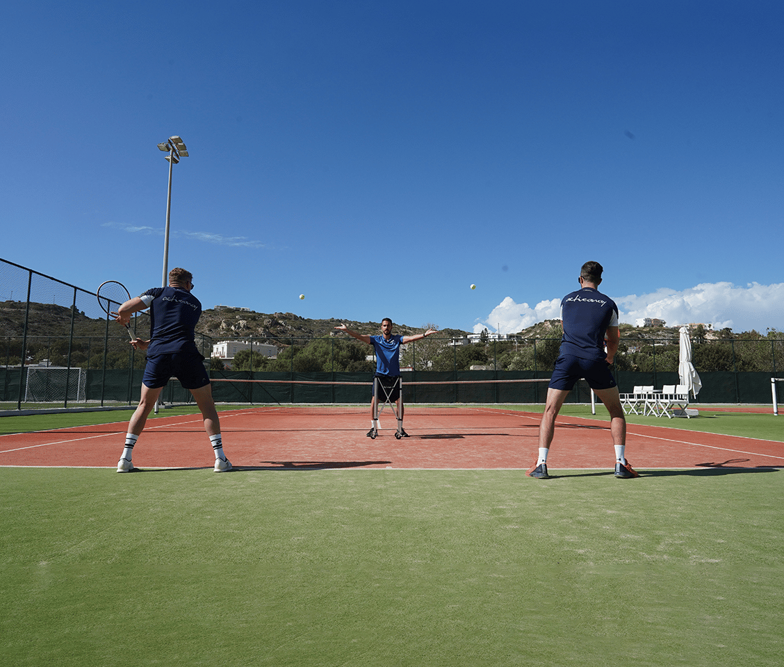 Outdoor tennis coaching session with a coach serving to two players on a clay court under a clear blue sky.