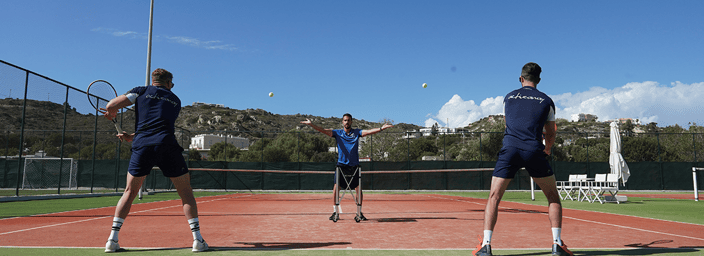 Outdoor tennis coaching session with a coach serving to two players on a clay court under a clear blue sky.