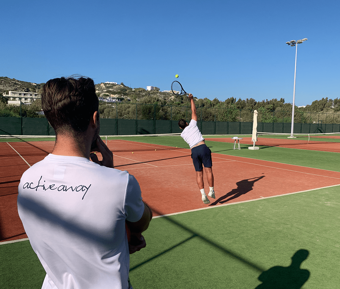 Tennis coach observing a player's serve on an outdoor court, with the coach's shirt displaying 'achieve away.'