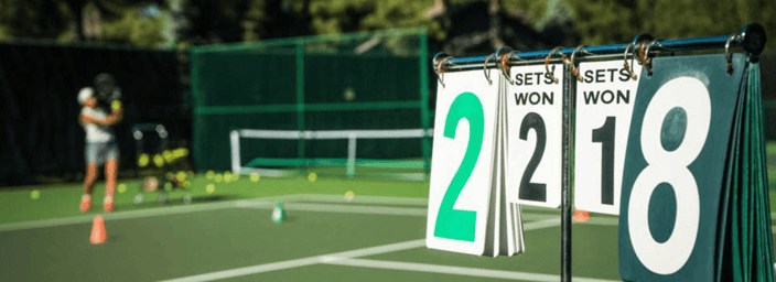 Tennis court with a scorecard showing 2-2 sets, person practising in the background with tennis balls on the court.