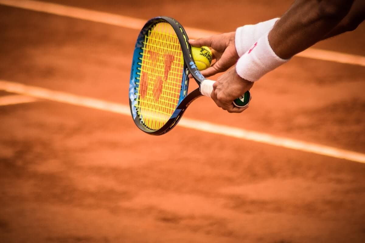 Close-up of a tennis player's hand holding a tennis ball and racquet, ready to serve on a clay court.