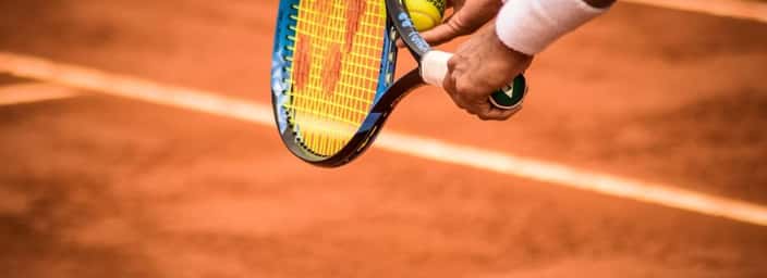 Close-up of a tennis player's hand holding a tennis ball and racquet, ready to serve on a clay court.