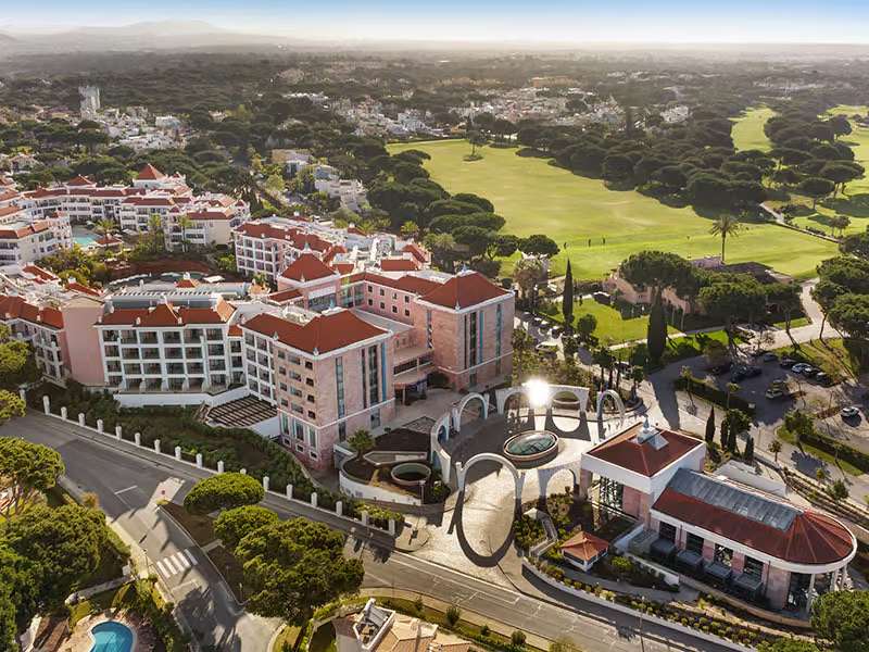 Aerial view of Hilton Vilamoura As Cascatas Golf Resort & Spa, featuring the hotel buildings with red-tiled roofs and an adjacent golf course in lush green surroundings.