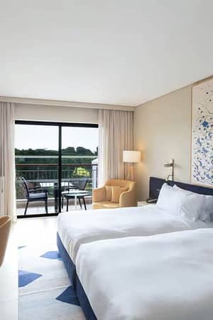 Hilton Vilamoura hotel room with twin beds, abstract wall art, a desk, TV, and a balcony overlooking greenery.