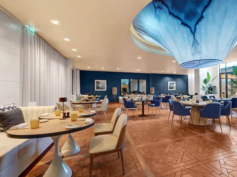 Indoor dining area at Hilton Vilamoura showcasing elegant decor with blue and beige chairs, parquet flooring, and a unique blue wave ceiling fixture.