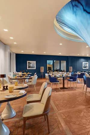 Indoor dining area at Hilton Vilamoura showcasing elegant decor with blue and beige chairs, parquet flooring, and a unique blue wave ceiling fixture.
