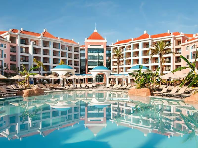 Wide shot of Hilton Vilamoura resort, featuring the hotel exterior with a grand swimming pool and sun loungers in the foreground.