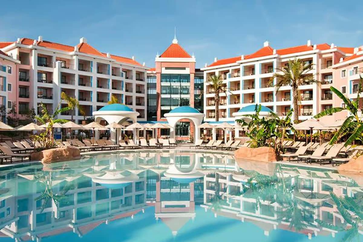 Wide shot of Hilton Vilamoura resort, featuring the hotel exterior with a grand swimming pool and sun loungers in the foreground.