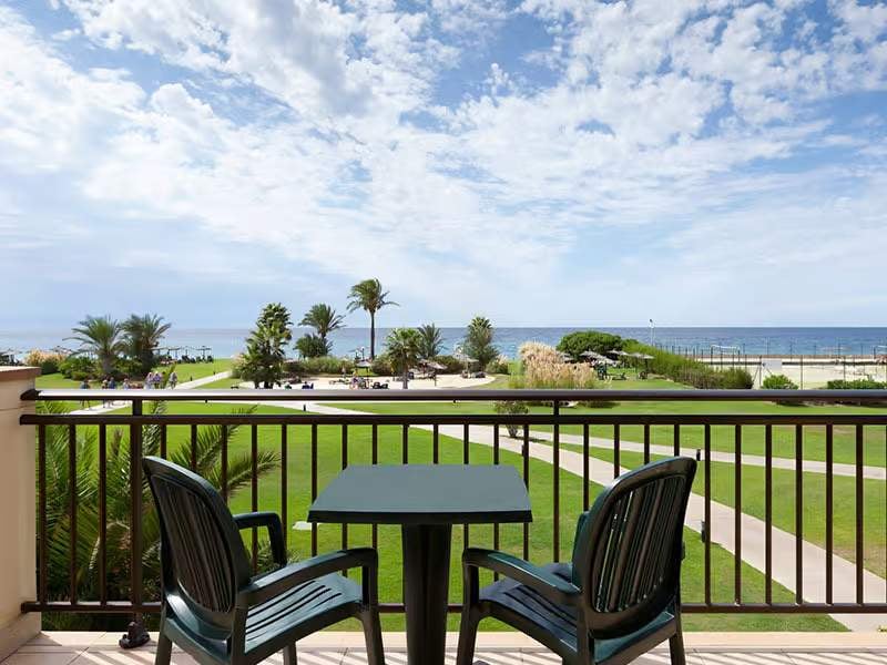 Hotel balcony view with two chairs and a table looking out onto gardens and the sea under a blue sky with clouds.