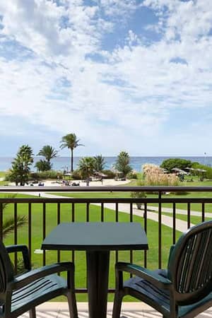 Hotel balcony view with two chairs and a table looking out onto gardens and the sea under a blue sky with clouds.