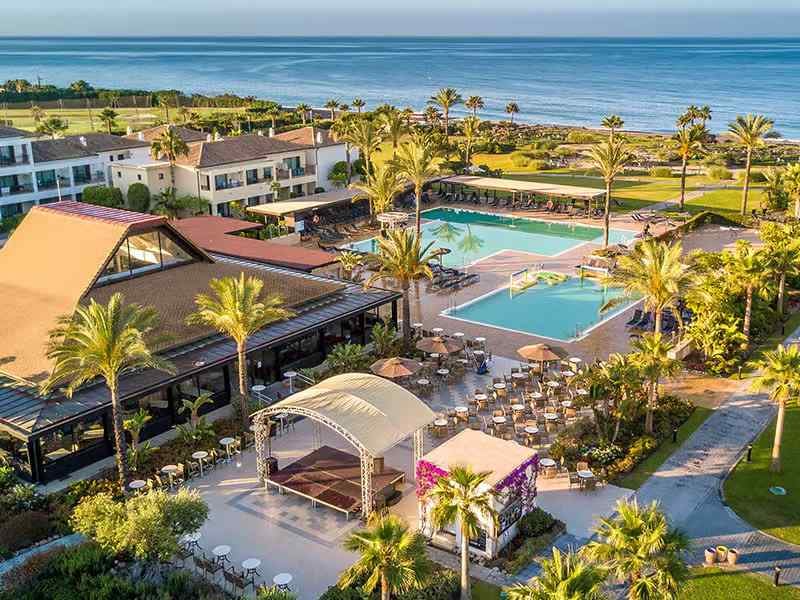 Aerial view of Hotel Impressive's pool area with two pools, sun loungers, palm trees, lush gardens, and the ocean in the background.