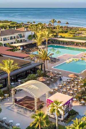 Aerial view of Hotel Impressive's pool area with two pools, sun loungers, palm trees, lush gardens, and the ocean in the background.