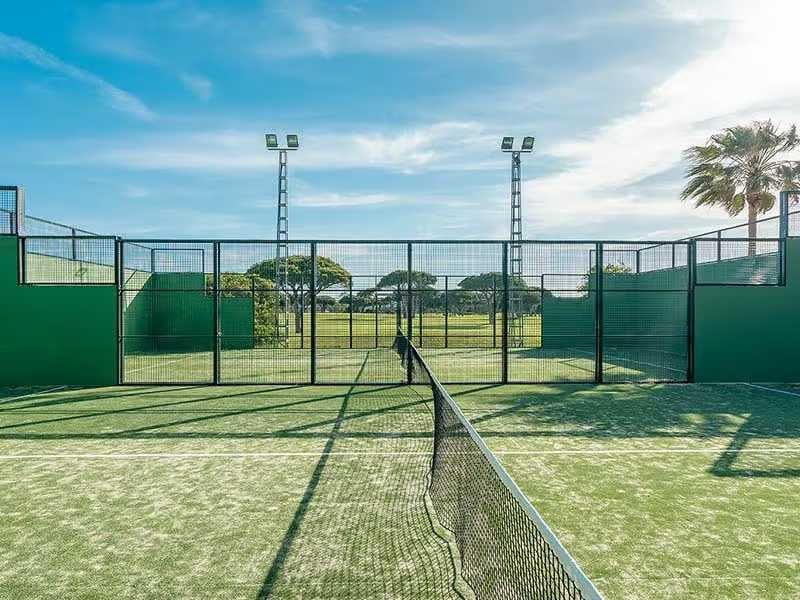 Padel tennis court with green artificial grass and metal fencing at Hotel Vincci, with palm trees and a blue sky in the background.