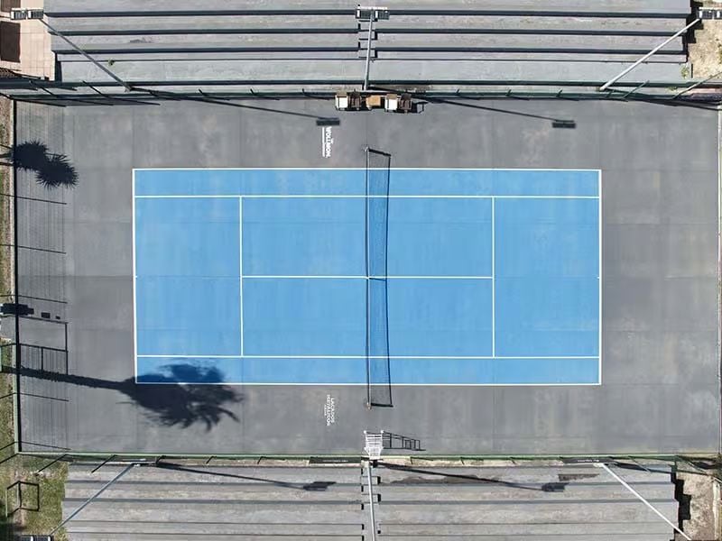 Aerial view of a vibrant blue tennis court at Hotel Vincci, with surrounding stands and palm trees casting shadows.