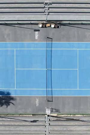 Aerial view of a vibrant blue tennis court at Hotel Vincci, with surrounding stands and palm trees casting shadows.