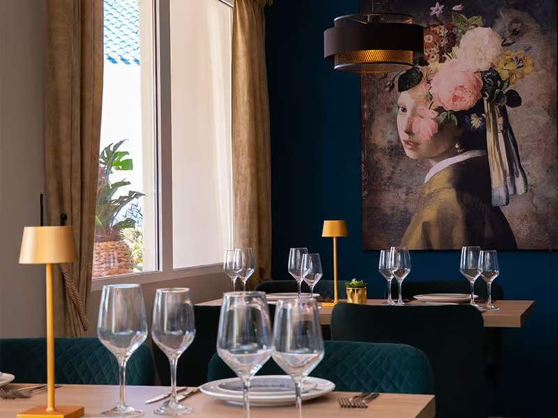Chic dining area with elegantly set tables, golden lamps, and a wall art piece of a woman with flowers in her hair.
