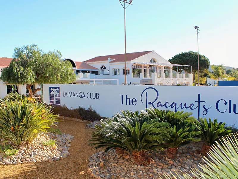 La Manga Club hotel exterior with garden and The Racquets Club sign, featuring palm trees and bright sky.