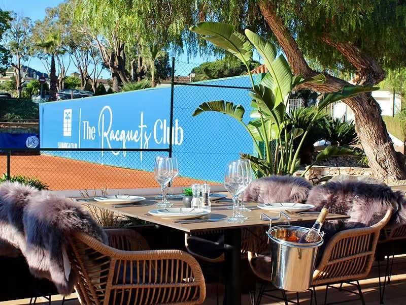 Outdoor dining area at The Racquets Club in La Manga, with table settings, fur-lined chairs, and a view of the tennis court and plants.