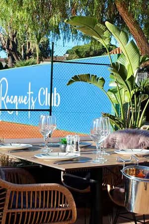 Outdoor dining area at The Racquets Club in La Manga, with table settings, fur-lined chairs, and a view of the tennis court and plants.