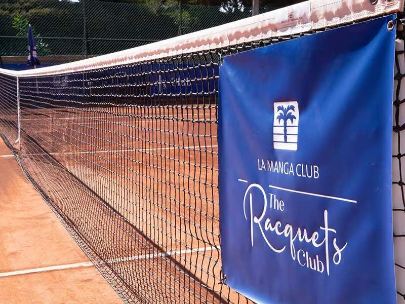 Close-up of a tennis net on a clay court at La Manga Club's The Racquets Club, with a blue banner displaying the club's logo.