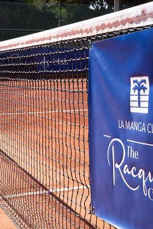Close-up of a tennis net on a clay court at La Manga Club's The Racquets Club, with a blue banner displaying the club's logo.