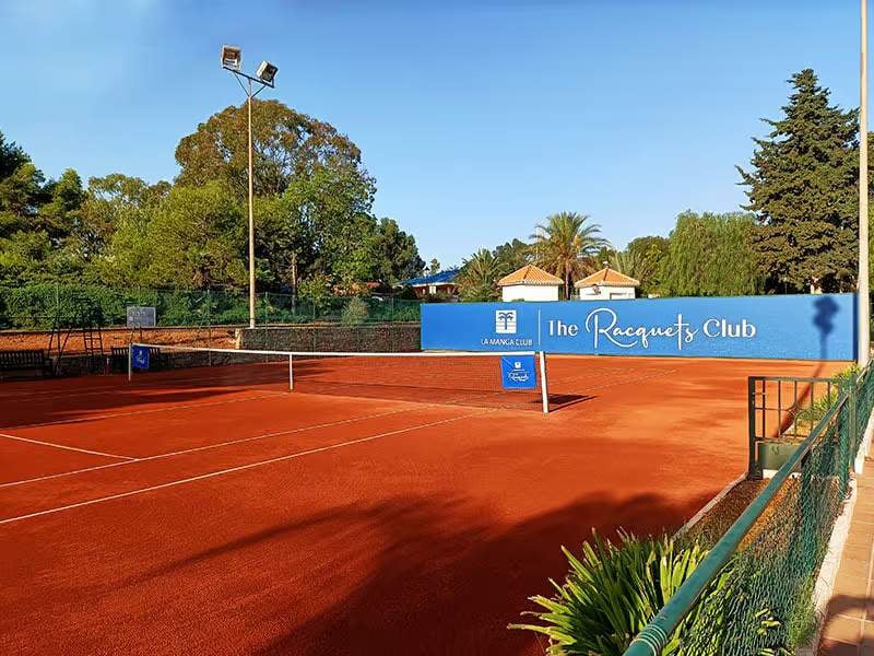 Wide shot of a clay tennis court at The Racquets Club, La Manga, with trees and a clear sky in the background.
