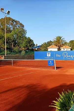 Wide shot of a clay tennis court at The Racquets Club, La Manga, with trees and a clear sky in the background.
