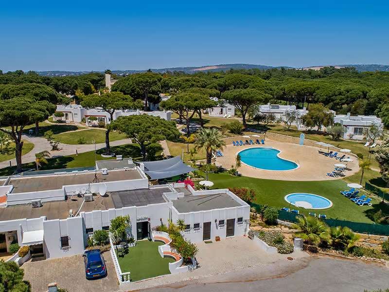 Aerial view of Prado Vilamoura with hotel villas, two swimming pools with sun loungers, amidst lush greenery and pine trees.