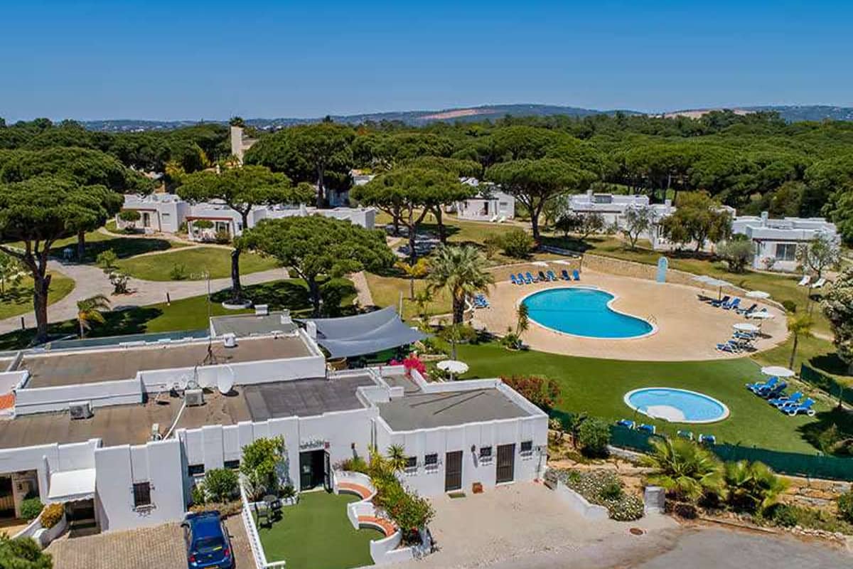Aerial view of Prado Vilamoura with hotel villas, two swimming pools with sun loungers, amidst lush greenery and pine trees.