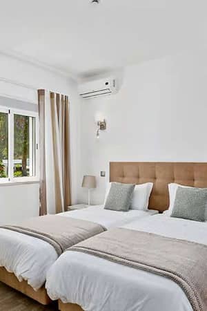 Hotel room at Prado Vilamoura with twin beds, modern decor, wooden headboard, ambient lighting, and a window with beige curtains.