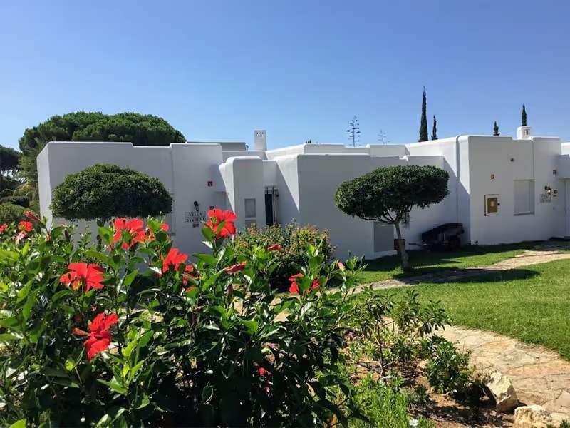 Prado Vilamoura Gallery Hotel Villas exterior with white geometric buildings, lush lawns, and red hibiscus flowers.
